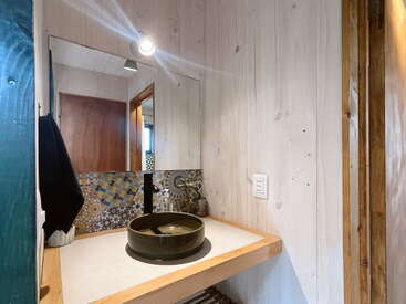Dieses gemütliche Badezimmer verfügt über ein rundes grünes Waschbecken, Holzwände, einen großen Spiegel, einen eklektischen Mosaikaufsatz, einen Strahler, ein Handtuch und eine Steckdose an der Wand.