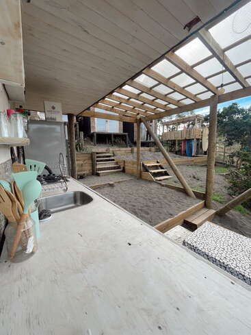 Dieses Bild zeigt eine rustikale Außenküche mit einer hölzernen Arbeitsplatte, einer Spüle, Utensilien und einem Kühlschrank. Die überdachte Terrasse blickt auf einen sandigen Hof und eine Holztreppe.
