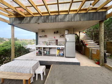 Dieses Bild zeigt eine rustikale Außenküche mit einem überdachten Dach, einem Kühlschrank, einer Spüle, offenen Regalen, einem Picknicktisch, Holzbänken, Sandboden und Stufen, die nach oben führen.
