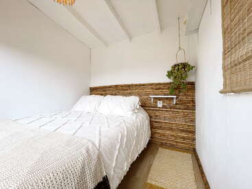 Ein gemütliches, minimalistisches Schlafzimmer mit weißen Wänden, einem Kopfteil aus Bambus, weicher Bettwäsche, einer gehäkelten Decke, einem gewebten Teppich, einer Hängepflanze und einem Bambusfensterschirm.