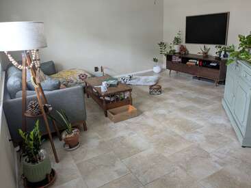 Ein gemütliches Wohnzimmer mit einem grauen Sofa, gemusterten Kissen, ein paar Pflanzen, einem Couchtisch aus Holz, einem Fernseher, einem grünen Schrank und Fliesenboden. Helle, minimalistische Einrichtung.