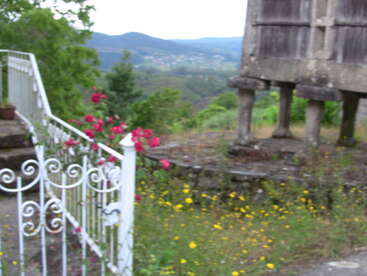 A imagem mostra uma cena serena ao ar livre com uma grade branca, uma estrutura de pedra e flores vibrantes, em um pitoresco cenário montanhoso.