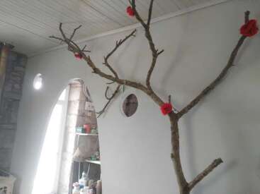 A imagem retrata um cômodo com um galho de árvore nu adornado com flores vermelhas, contra uma parede e um teto brancos, com uma janela e prateleiras visíveis ao fundo.