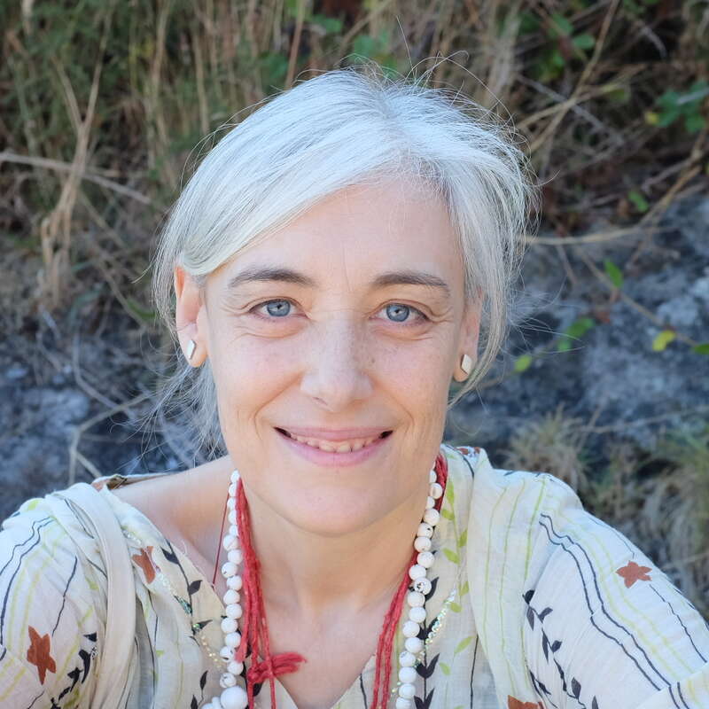 A imagem mostra uma mulher com cabelos brancos, olhos azuis e blusa branca, sorrindo para a câmera, com um colar e brincos, em frente a um fundo natural ao ar livre.