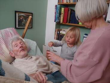 Ein älterer Mann liegt mit einer Decke im Bett, assistiert von einer älteren Frau und einem kleinen Kind. Das Zimmer ist gemütlich, mit Bücherregalen und gerahmten Bildern.