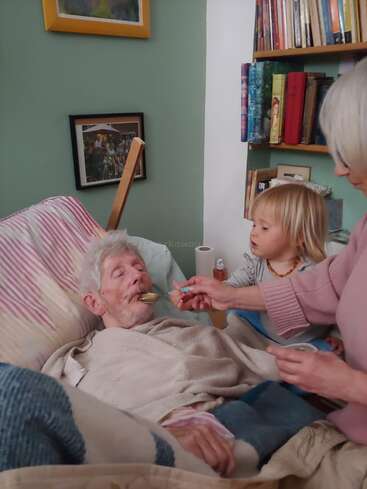 Ein älterer Mann liegt im Bett und wird von einer Frau sanft gefüttert, während ein kleines Kind aufmerksam zusieht. Bücher und gerahmte Fotos schmücken den gemütlichen Raum.