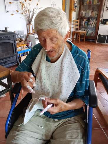 The image shows an elderly man sitting in a wheelchair, wearing a bib and holding a piece of paper, with a warm and cozy living room in the background.