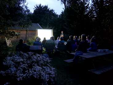 Un groupe de personnes est assis en plein air, la nuit, et regarde un écran de projection très lumineux. La scène est entourée d'arbres et de fleurs, créant une atmosphère chaleureuse.