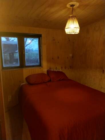 Cette petite chambre confortable présente des murs en bois, un lit double avec des draps rouges, une fenêtre avec vue sur les arbres et un luminaire chaud et tressé suspendu au-dessus.