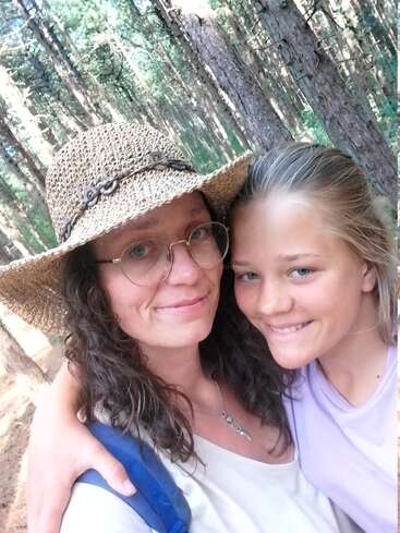 Une femme portant un chapeau de paille et des lunettes serre dans ses bras une jeune fille souriante. Elles se tiennent l'une à côté de l'autre dans une forêt ensoleillée remplie de grands arbres.