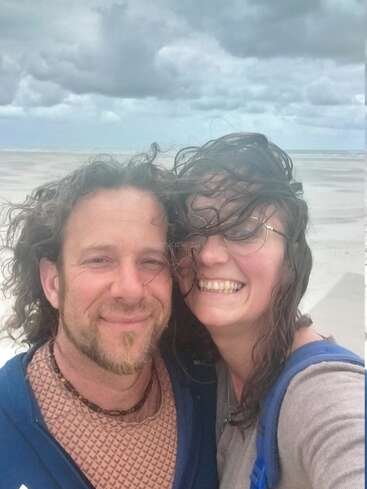 Un couple souriant prend un selfie sur une plage venteuse. Leurs cheveux sont ébouriffés par le vent et un ciel nuageux s'étend sur le rivage vide et sablonneux derrière eux.