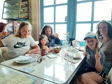 Une famille de cinq personnes, souriantes et faisant des signes de paix, est assise à une table de restaurant avec de la nourriture et des boissons, profitant d'un repas joyeux près de fenêtres lumineuses.