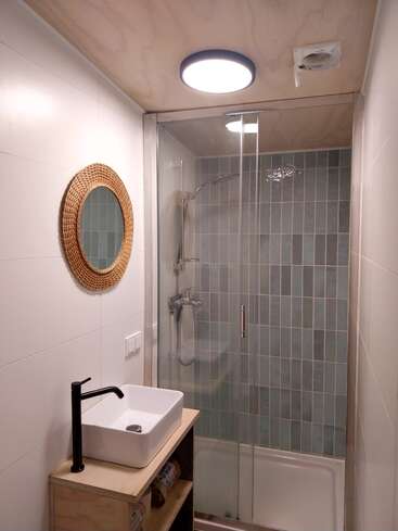 Une petite salle de bains moderne avec une douche vitrée, un mur carrelé bleu, un miroir rond tissé, un lavabo, un robinet noir et un meuble en bois sous un éclairage doux au plafond.
