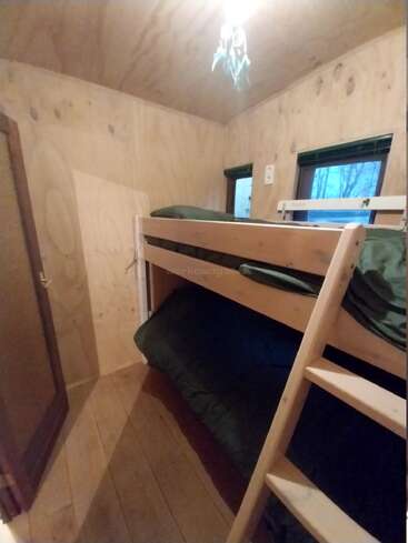 Cette petite chambre confortable présente des murs et un plafond en bois, un lit superposé avec des draps verts, deux fenêtres pour la lumière naturelle et un simple plafonnier.