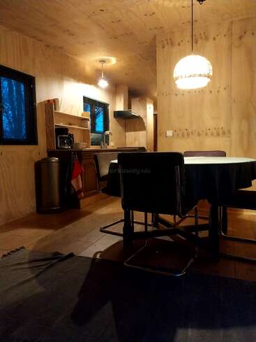 Une cuisine et une salle à manger confortables avec des murs et un plafond en bois, des chaises noires modernes, une table ronde, un éclairage chaleureux, des étagères ouvertes et une fenêtre laissant apparaître un ciel bleu.