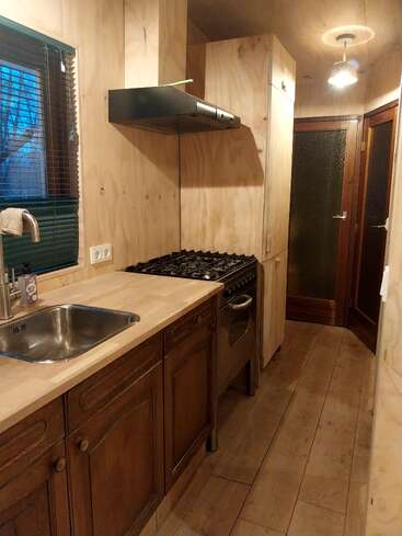 Cette cuisine est équipée de boiseries, d'une cuisinière à gaz, d'un évier en acier inoxydable, d'armoires en bois et d'un éclairage moderne. Une fenêtre avec stores apporte la lumière naturelle à l'intérieur.