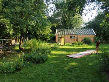 Une arrière-cour verdoyante avec une cabane en bois, un toboggan pour enfants, des chaises d'extérieur, des plantes de jardin, des arbres matures et une personne debout près d'un tapis rectangulaire. Atmosphère paisible.