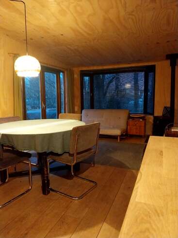 Gemütliches Esszimmer aus Holz mit rundem Tisch, vier Stühlen, Hängelampe, großen Fenstern, grüner Tischdecke, Futonsofa, Beistelltisch und warmem Tageslicht.