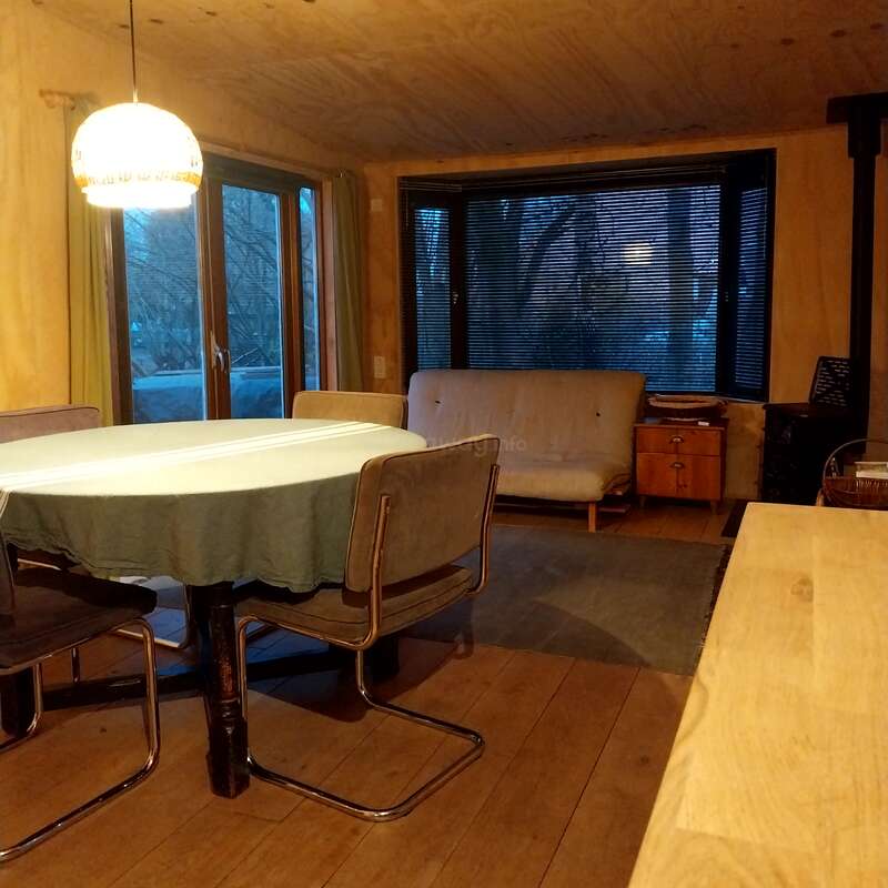 Salle à manger confortable en bois avec une table ronde, quatre chaises, une lampe suspendue, de grandes fenêtres, une nappe verte, un canapé futon, une table d'appoint et un éclairage naturel chaleureux.