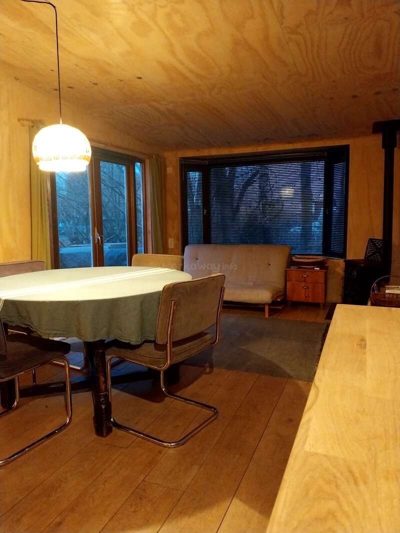 Salle à manger confortable en bois avec une table ronde, quatre chaises, une lampe suspendue, de grandes fenêtres, une nappe verte, un canapé futon, une table d'appoint et un éclairage naturel chaleureux.