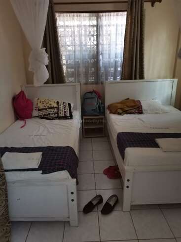 Cette chambre confortable comprend deux lits simples avec des draps blancs, des oreillers colorés, une fenêtre avec des rideaux, des sacs à dos placés entre les lits et des chaussures bien rangées sur le sol.