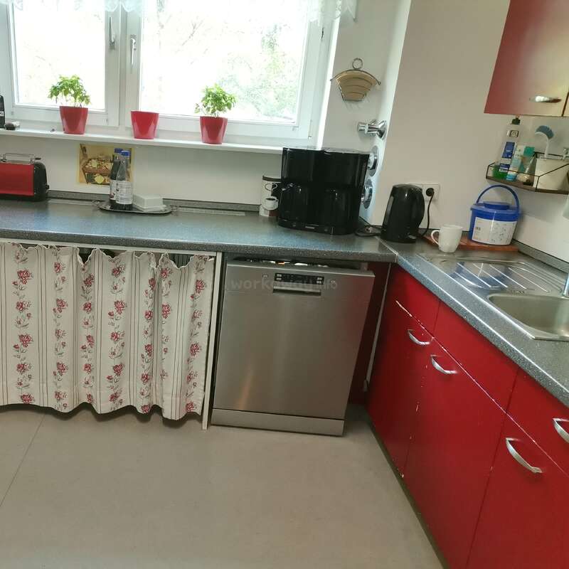 Une cuisine confortable avec des armoires rouges, des rideaux à fleurs, un lave-vaisselle argenté, une cafetière, une bouilloire, un grille-pain, des herbes en pot sur le rebord de la fenêtre et divers articles de cuisine sur les comptoirs.