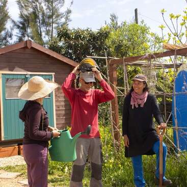 Three people stand outside near a wooden shed, engaged in conversation. One holds a watering can, another adjusts a helmet, and the third leans on a shovel.