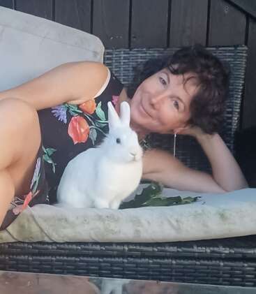 Une femme vêtue d'une robe à fleurs est allongée sur un banc rembourré en plein air ; elle sourit, détendue, aux côtés d'un lapin blanc tout doux. La scène dégage une atmosphère calme et chaleureuse.