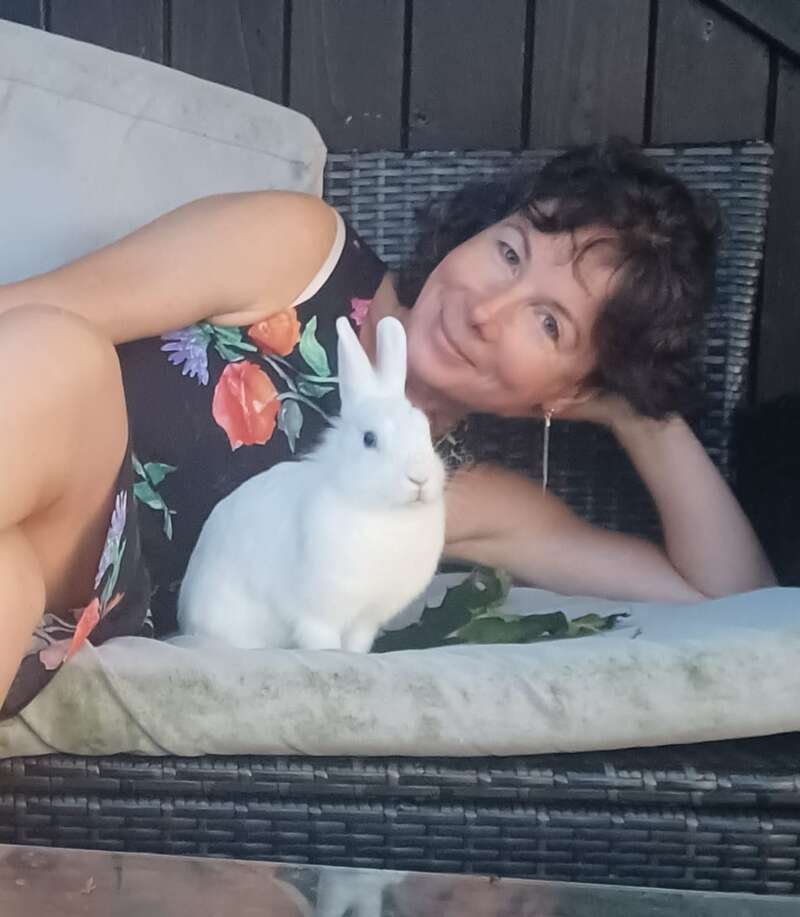 Une femme vêtue d'une robe à fleurs est allongée sur un banc rembourré en plein air ; elle sourit, détendue, aux côtés d'un lapin blanc tout doux. La scène dégage une atmosphère calme et chaleureuse.