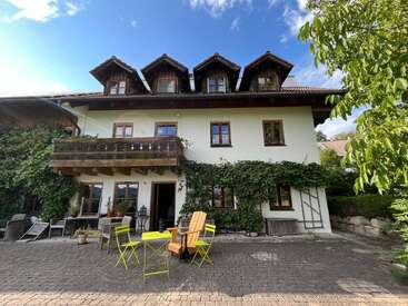 Ein charmantes, zweistöckiges Haus mit Efeu an den Wänden, einem Holzbalkon und Dachgauben. Leuchtend gelbe Gartenmöbel und üppiges Grün schaffen eine einladende Atmosphäre.