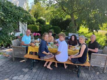 Sept personnes prennent leur repas ensemble à une table en plein air, sous un arbre, entourées de verdure et baignées par la lumière du soleil, partageant leur repas et discutant dans une atmosphère détendue.
