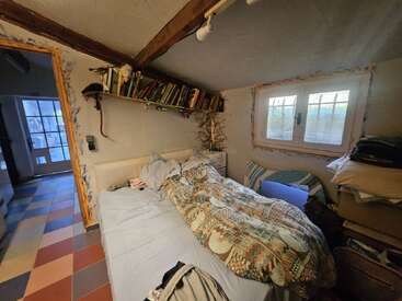 Une chambre confortable et encombrée comprend un lit simple avec une literie à motifs, des étagères au-dessus, des poutres en bois, une fenêtre et un sol carrelé coloré, menant à un couloir.