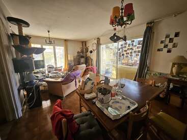Un salon encombré avec une décoration éclectique, un arbre à chat, des meubles éparpillés, une table à manger avec des objets, de grandes fenêtres, une lumière vive, des photos aux murs et un lustre coloré suspendu.