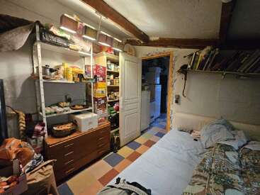 Une pièce confortable et encombrée, avec des étagères remplies de nourriture et de provisions, un lit simple, des livres, un sol carrelé coloré et une porte ouverte menant à une autre pièce.