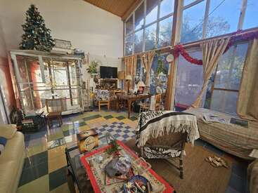 Un salon confortable et lumineux avec de grandes fenêtres, des décorations de Noël, une cage à oiseaux, un sol carrelé coloré, des meubles assortis, des plantes et une table basse encombrée.
