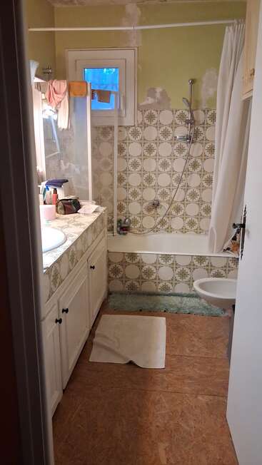 Cette image montre une vieille salle de bain avec des carreaux à motifs, une baignoire, une douche, une petite fenêtre, un lavabo encombré, des armoires blanches, des murs inachevés et un sol en bois.