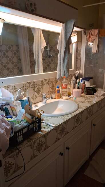 Une salle de bain encombrée avec un lavabo, un miroir, des serviettes, des produits d'hygiène personnelle, des articles de toilette, une caisse d'objets et un éclairage zénithal lumineux.
