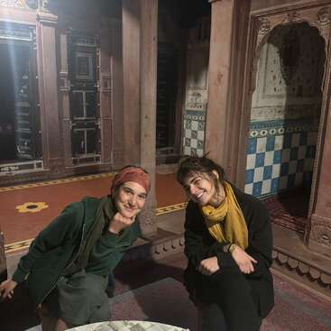 The image depicts two women posing in a traditional Indian building, with ornate carvings and colorful tiles, conveying a sense of cultural richness and heritage.