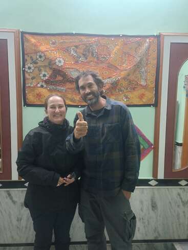 A smiling man and woman stand indoors, posing in front of an ornate, colorful tapestry. The man gives a thumbs-up; both wear casual, dark-colored clothing.