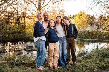Eine fünfköpfige Familie posiert gemeinsam im Freien in der Nähe eines Teiches, umgeben von Bäumen und sanftem Sonnenlicht. Sie sind lässig gekleidet, lächeln und zeigen starke Verbundenheit.