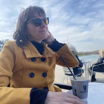 The image depicts a woman in a yellow coat sitting at a table with a cup of coffee, set against a serene outdoor backdrop featuring a body of water and a blue sky with clouds.