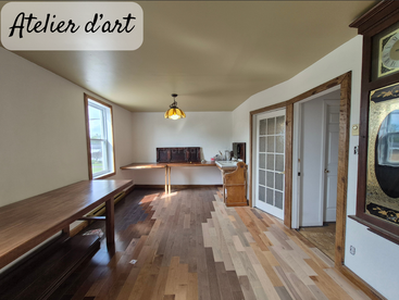 A bright, cozy art studio with wooden tables, parquet flooring, and vintage decor. Large windows let in light. Glass doors and a classic clock add charm.