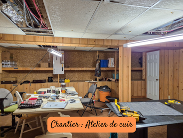This image shows a cluttered workshop under renovation, with exposed ceilings, various tools, worktables, chairs, and materials. The sign reads "Chantier : Atelier de cuir".