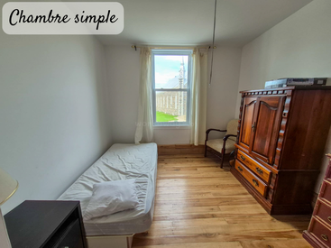 A small, simple bedroom with a single bed, wooden floor, vintage wardrobe, armchair, and window with white curtains. Sunlight brightens the cozy, minimalist space.