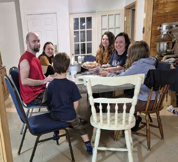 Six people, including children and adults, gather around a dining table, smiling and enjoying each other's company. The scene looks warm, casual, and friendly, indoors.