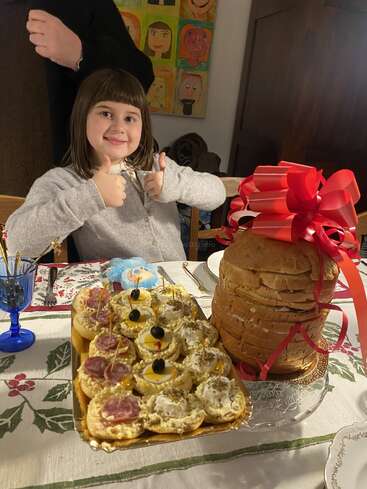 Une jeune fille souriante lève les deux pouces devant une table décorée de manière festive avec des amuse-gueules assortis et un grand pain de mie enveloppé d'un gros nœud rouge.