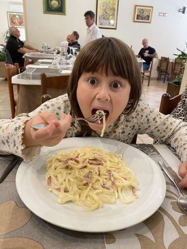 Un enfant aux yeux écarquillés mange avec enthousiasme des pâtes crémeuses au jambon dans un restaurant. D'autres personnes sont assises et dînent dans un cadre confortable et décoré.