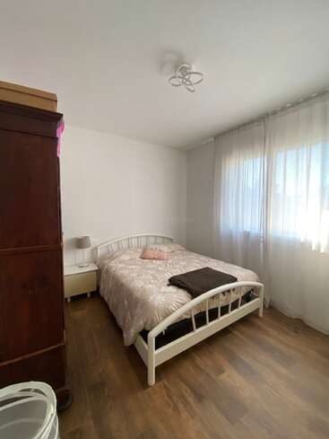 Das ist ein gemütliches Schlafzimmer mit einem weißen Metallbett, einer geblümten Bettdecke, einer braunen Decke, einem Holzboden, einem großen Kleiderschrank, einem Nachttisch, einer Lampe und transparenten Vorhängen, durch die das Sonnenlicht hereinfällt.