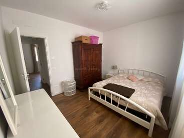 Das ist ein gemütliches Schlafzimmer mit einem Doppelbett, Holzboden, großem Kleiderschrank, Nachttisch, Lampe, Wäschekorb und schlichten weißen Wänden, die eine minimalistische Atmosphäre schaffen.