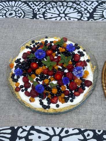 A delicious tart topped with whipped cream, fresh strawberries, blackcurrants, cloudberries, blue edible flowers, and mint leaves, displayed on a rustic linen tablecloth.